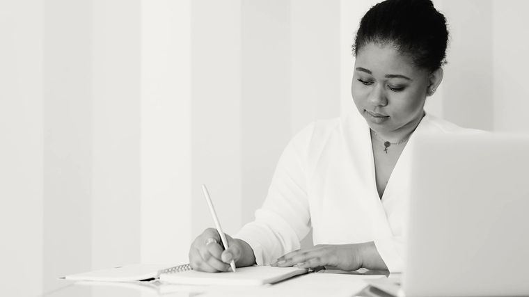A woman writing notes in a notebook beside her laptop.
